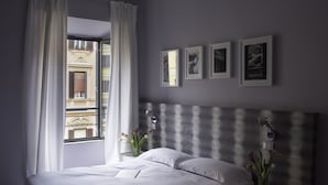 Une chambre à coucher avec un lit, une table de chevet, une lampe, un vase avec des fleurs et une fenêtre avec des rideaux.