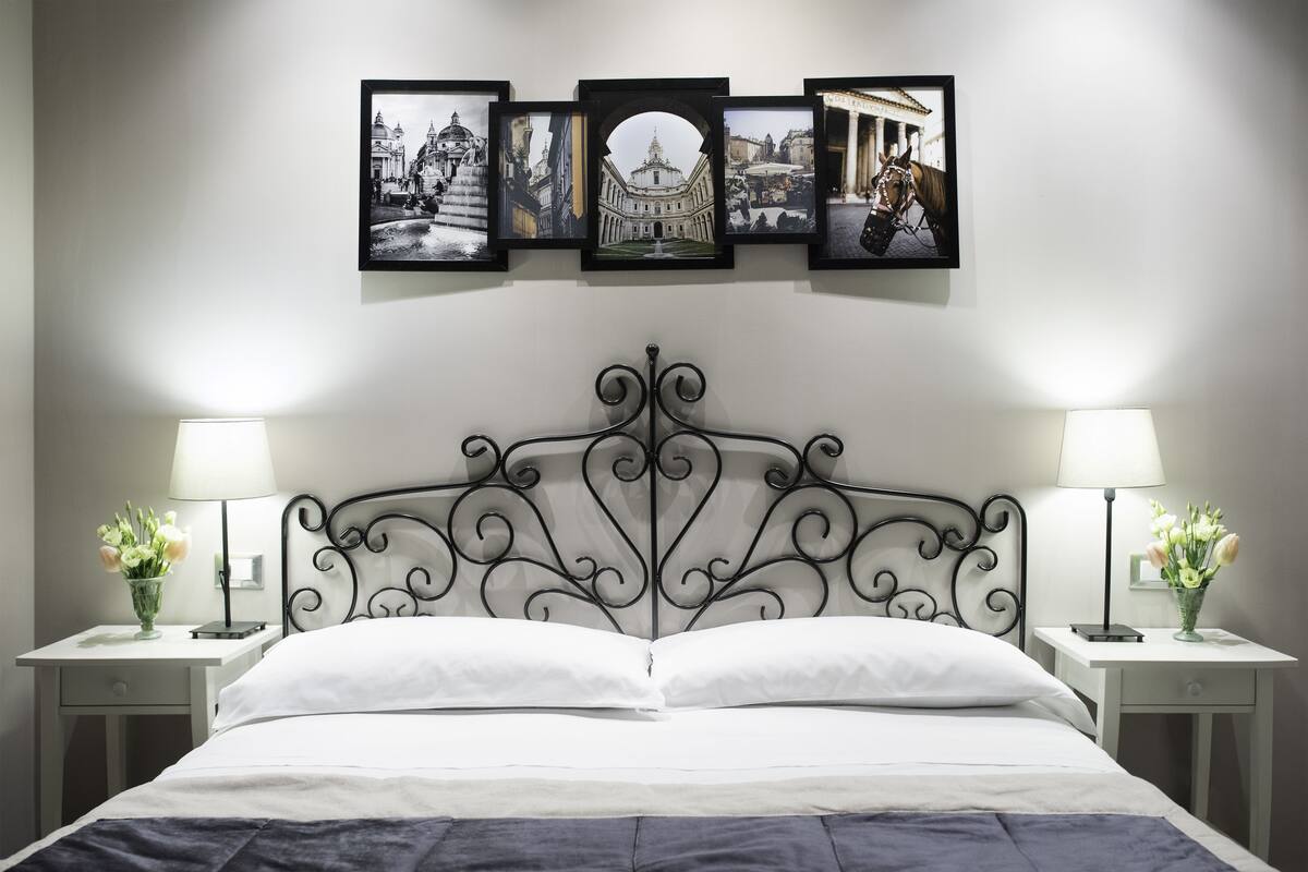 Una cama bien tendida con un cabecero decorativo, flanqueada por dos mesitas de noche con lámparas y jarrones. Encima de la cama, hay tres cuadros enmarcados en la pared.