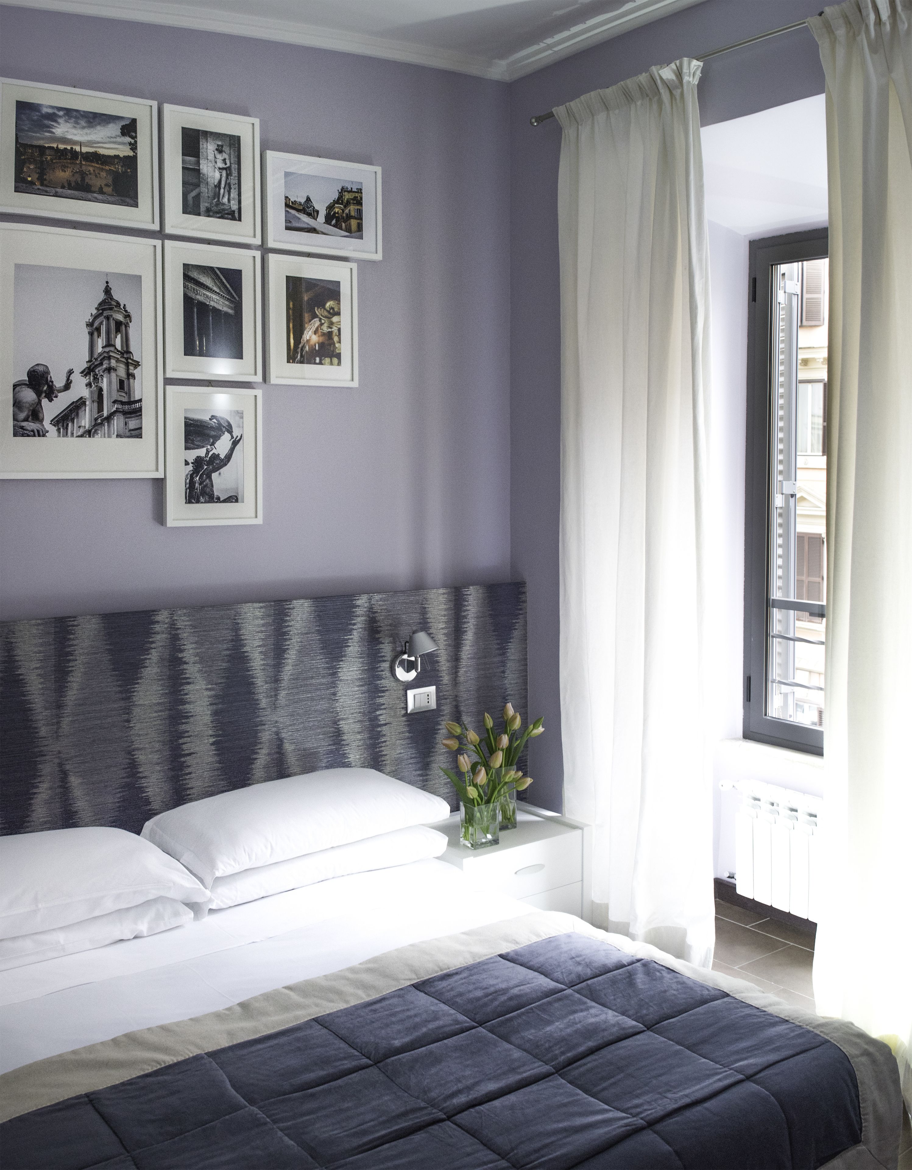 A bedroom with a bed, a headboard, a nightstand, a wall-mounted lamp, and a window with curtains.