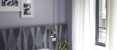 A bedroom with a bed, a headboard, a nightstand, a wall-mounted lamp, and a window with curtains.