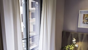 Une chambre avec une grande fenêtre, des rideaux blancs, une table de chevet ornée d’un vase de fleurs, un lit avec une tête de lit sombre et un lampadaire fixé au mur.