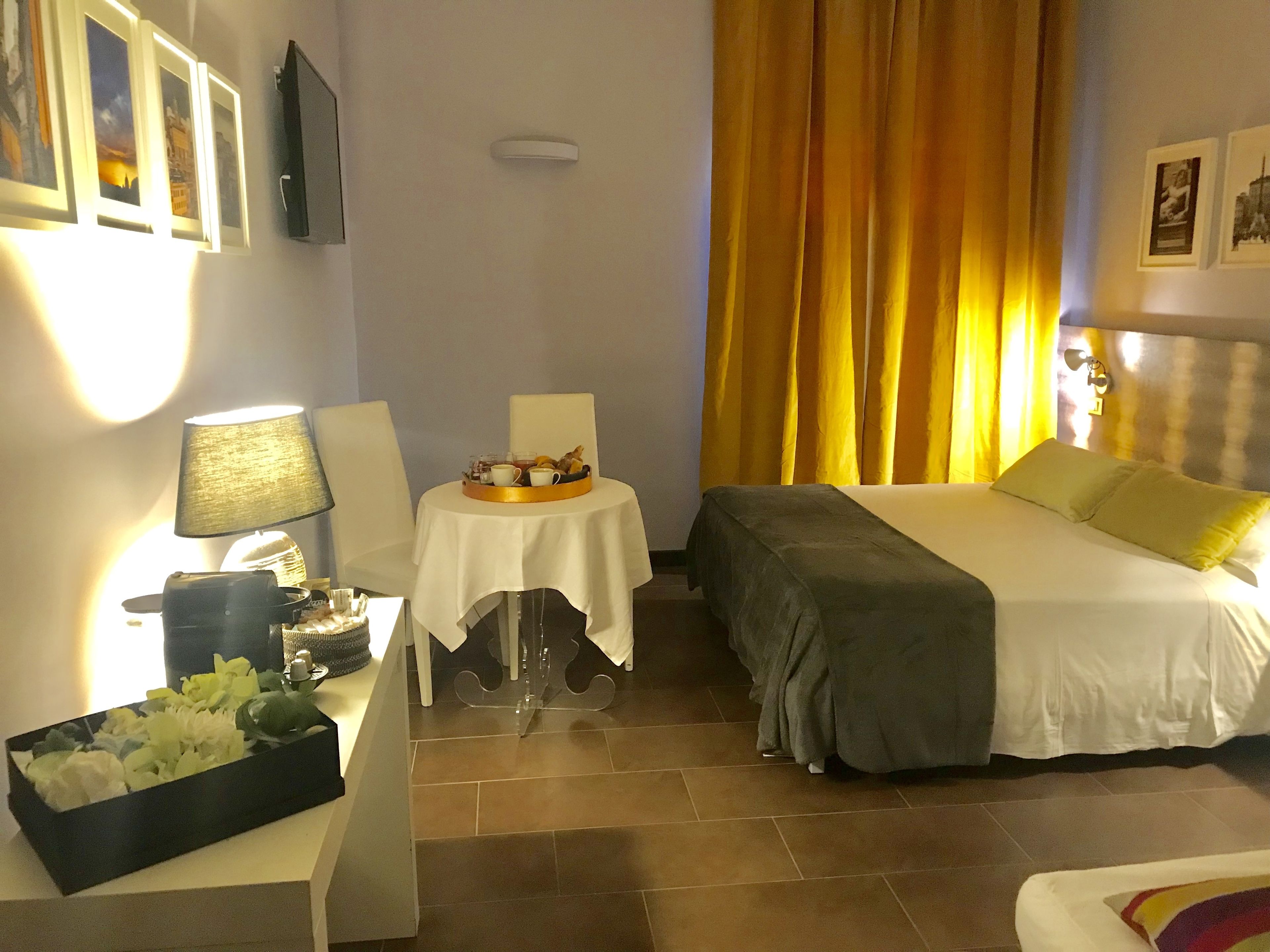 A hotel room with a bed, a small table with a tray of breakfast items, a lamp, and a flower arrangement.
