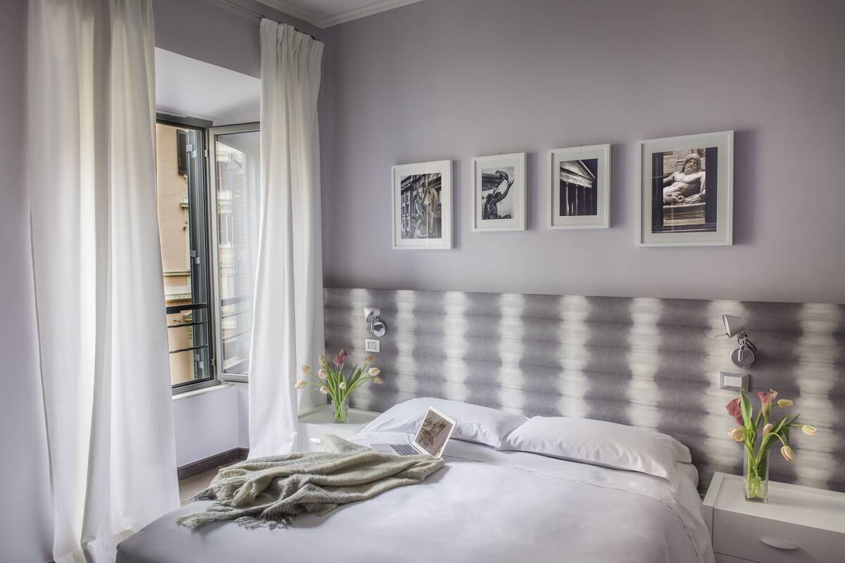 Un dormitorio moderno con una cama, una computadora portátil, una mesita de noche, un florero con flores y cuadros en la pared.