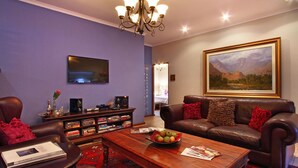 A living room with a brown leather sofa, a wooden coffee table with a fruit bowl, a bookshelf, a flat-screen TV, and a large framed landscape painting. - Apple Tree Guest House (Stellenbosch)