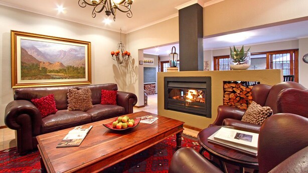 A cozy living room with a fireplace, leather sofas, a coffee table with apples, and a large framed landscape painting. - Apple Tree Guest House (Stellenbosch)