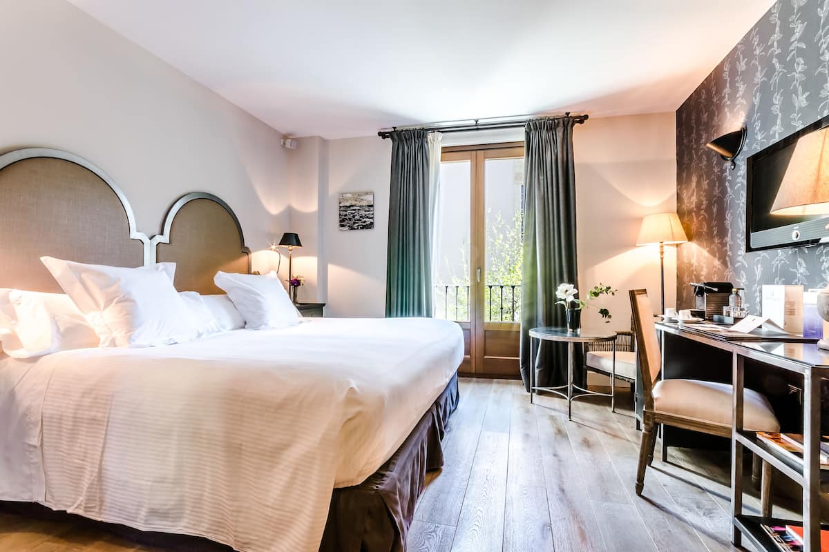 Ein Hotelzimmer mit einem großen Bett, einem Schreibtisch mit Stuhl, einem kleinen Tisch mit Lampe und Blick ins Freie durch ein Fenster.
