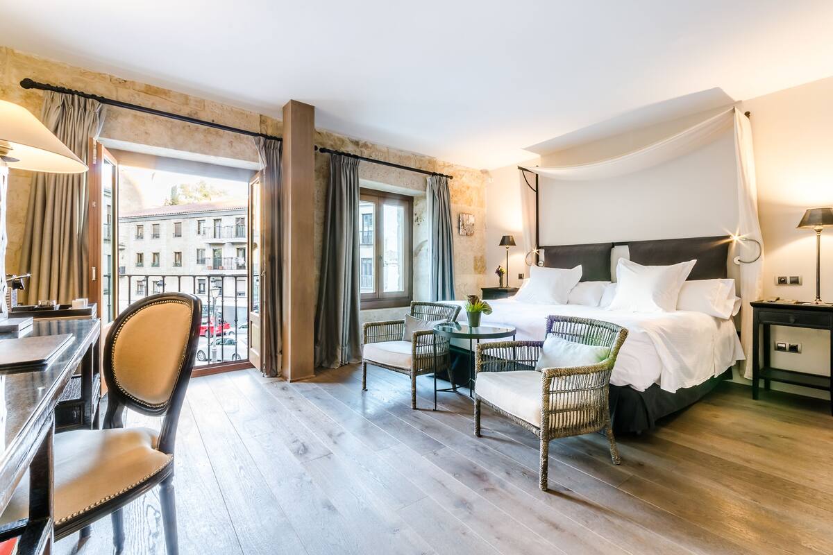 Ein Hotelzimmer mit einem großen Bett, zwei Korbsesseln, einem kleinen Tisch und Blick auf die Stadt durch ein großes Fenster.