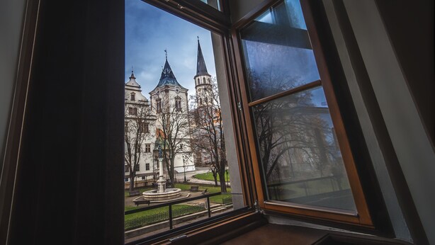 A view through a window of a building with two towers and a fountain in the courtyard.