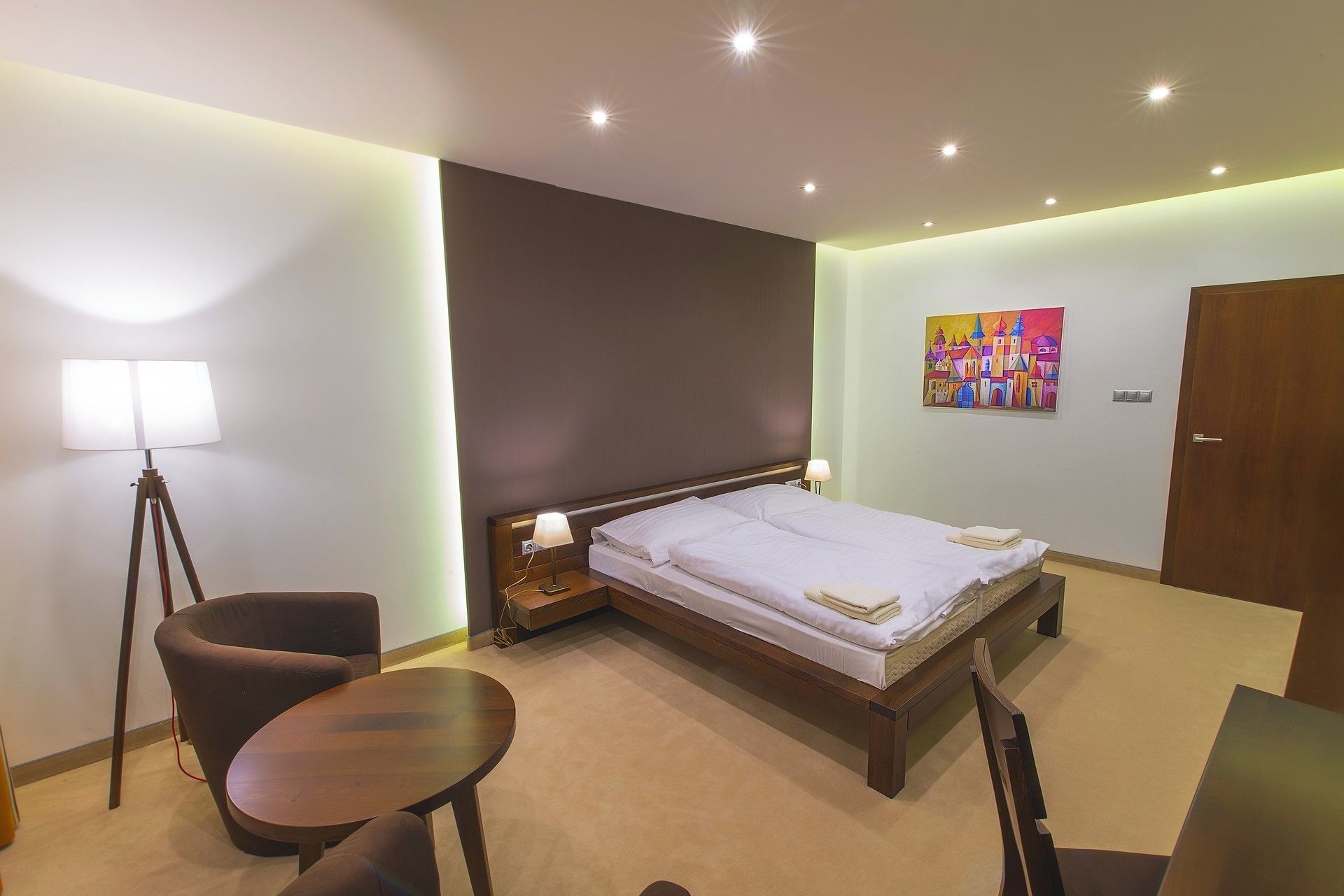 A hotel room with a bed, a chair, a small table, and a painting on the wall.