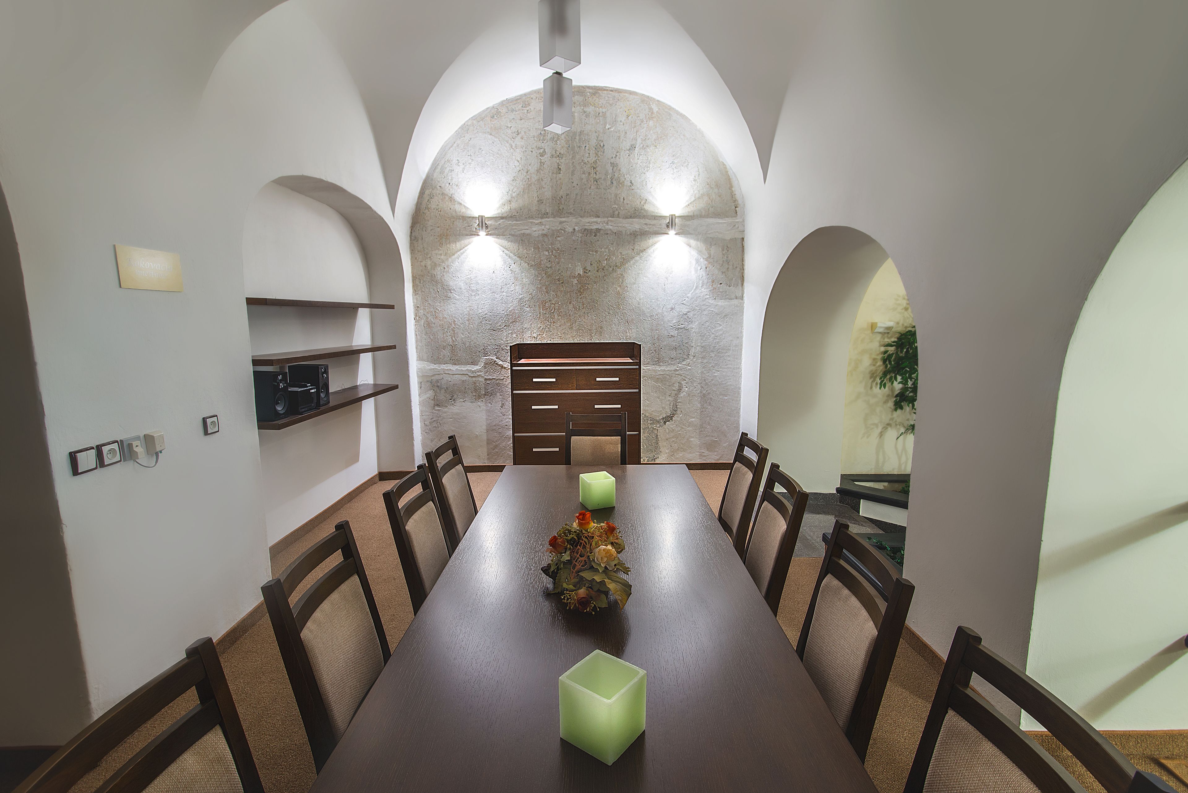 A dining area with a long table, chairs, a vase with flowers, and a stone wall with lights.