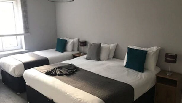Habitación de hotel con dos camas, una ventana con persianas, una mesita de noche con lámpara y un aplique de pared.