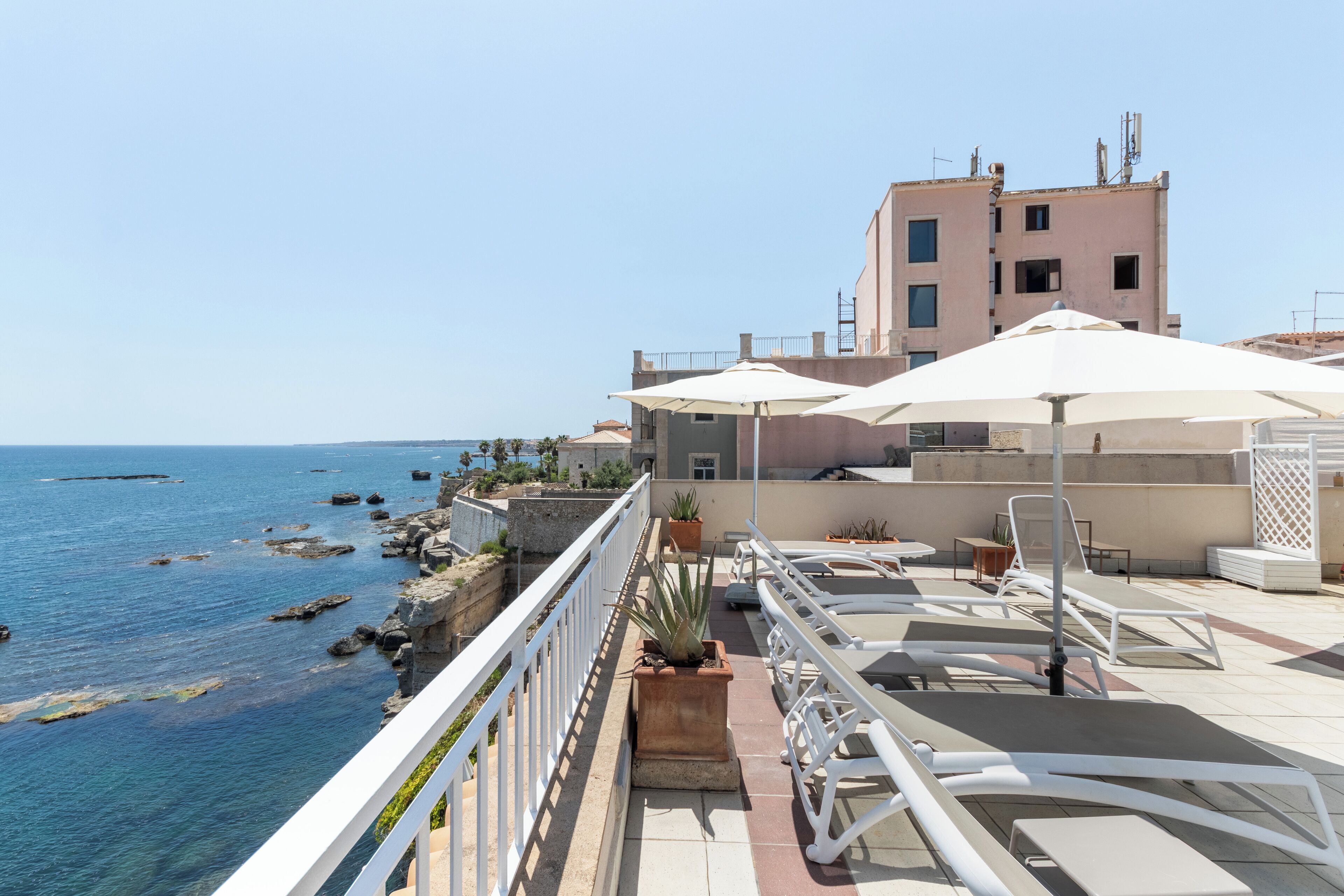 A seaside terrace with white railings, lounge chairs, and umbrellas overlooking the sea.