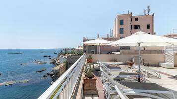 A seaside terrace with white railings, lounge chairs, and umbrellas overlooking the sea.