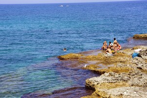 Escena costera con agua azul cristalina, costa rocosa y gente relajándose junto al mar.