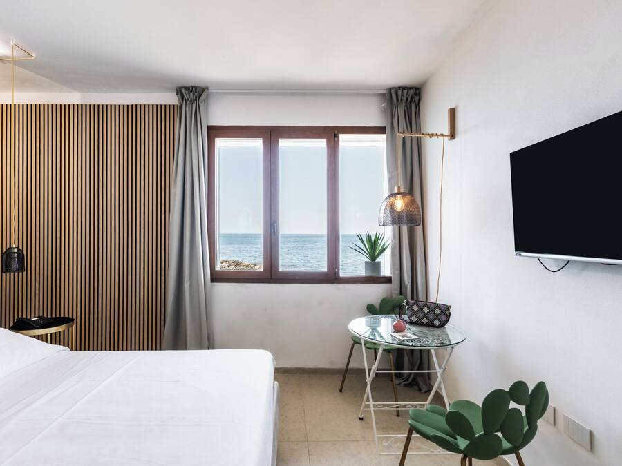 A hotel room with a bed, a small table with a chair, a TV mounted on the wall, and a window with a view of the sea.