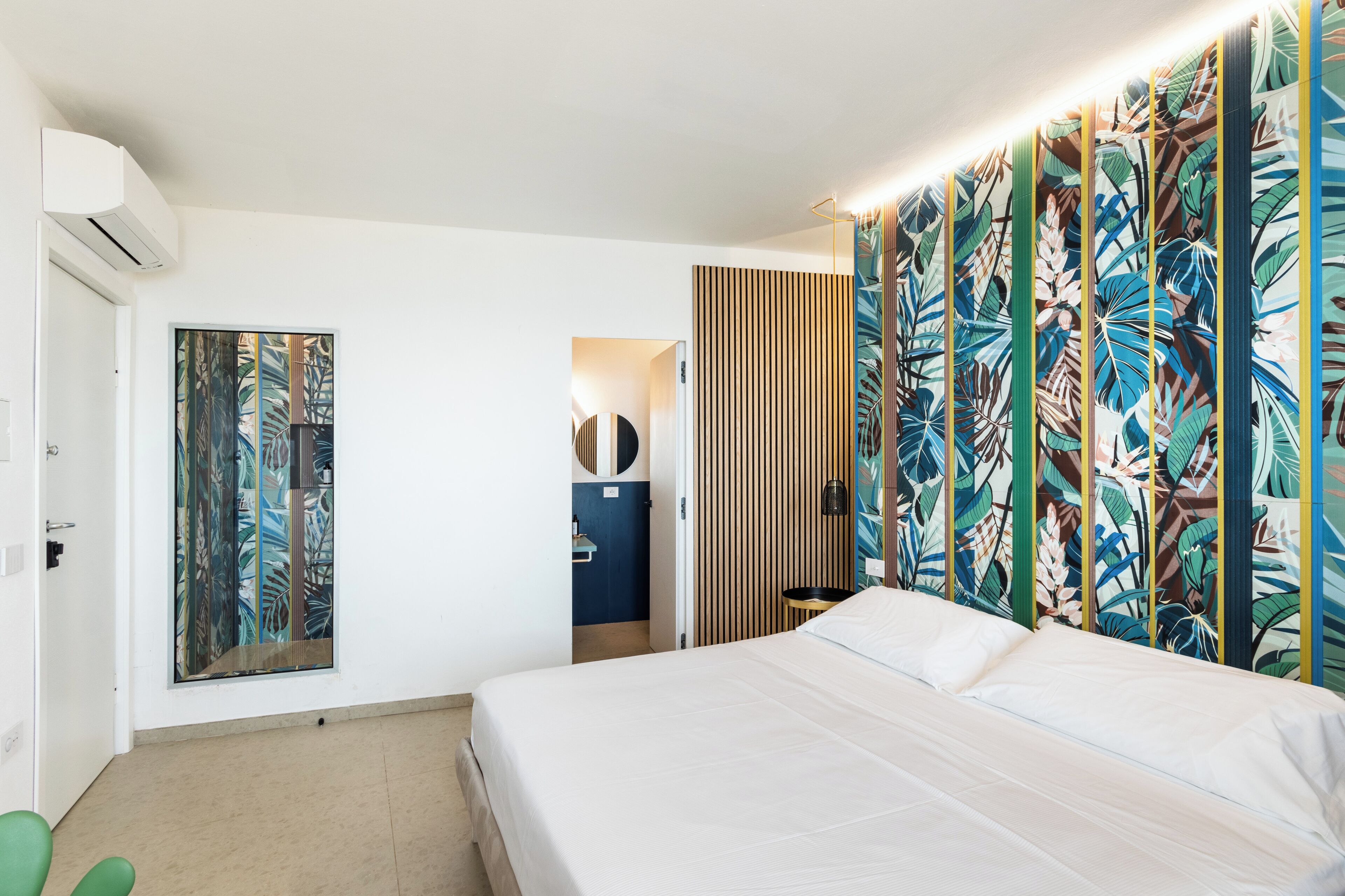 Une chambre d’hôtel avec un lit, un grand tableau aux motifs tropicaux, un mur recouvert de lambris en bois et une petite table avec une lampe.
