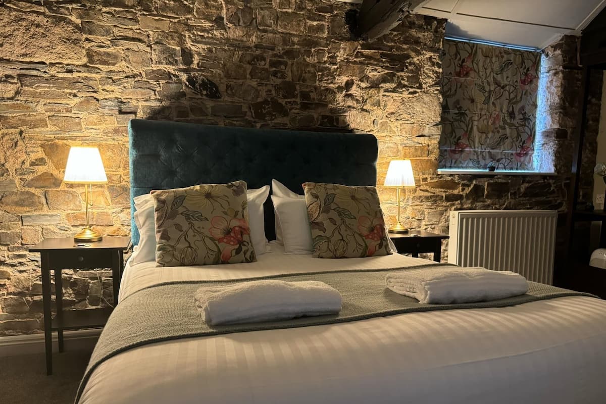 Quarto com parede de pedra, cama grande com almofadas florais, duas luminárias de cabeceira e um radiador.