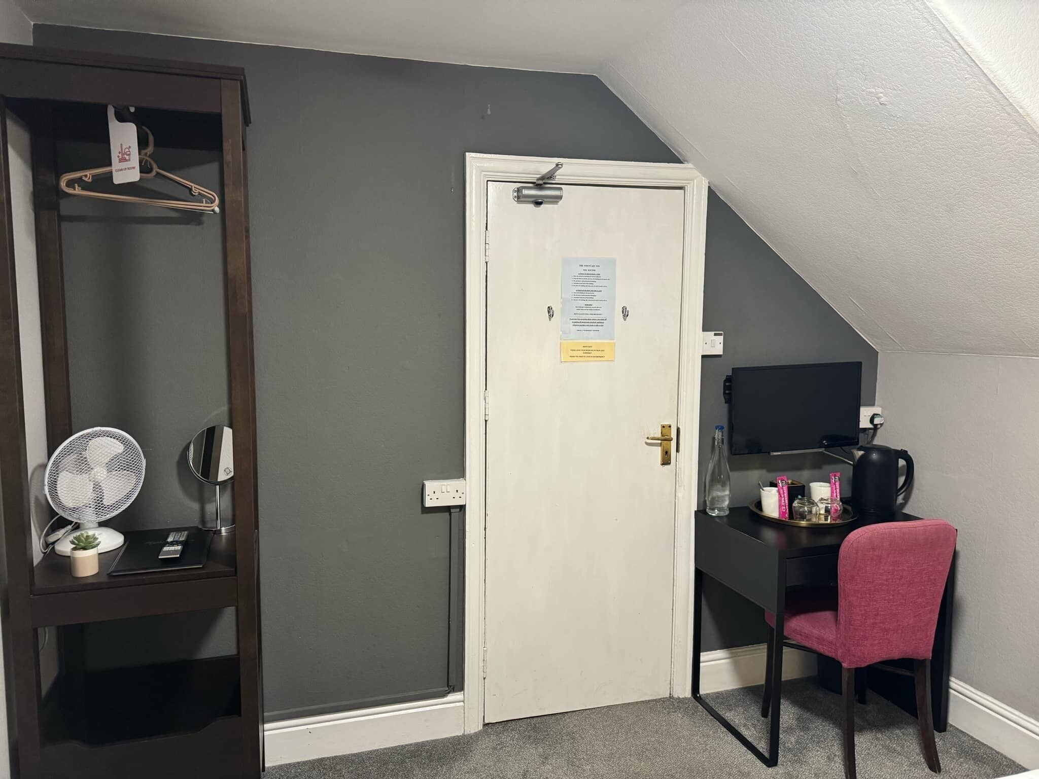 Una habitación con la puerta cerrada, un escritorio con una silla, una pequeña mesa con una planta, un ventilador y un perchero.