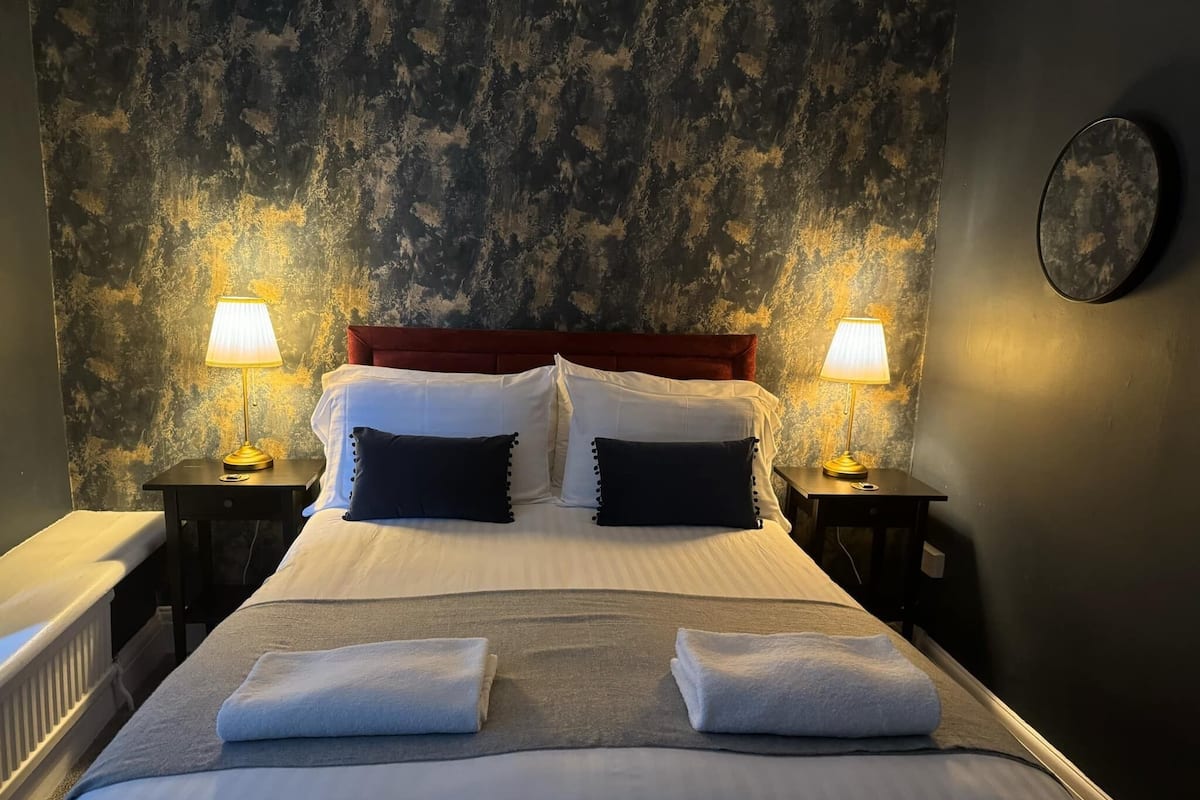 Uma cama bem arrumada com duas almofadas, duas mesas de cabeceira com luminárias e uma parede texturizada atrás da cama.