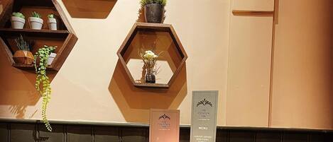 L’intérieur d’un restaurant présente des étagères hexagonales où sont exposées des plantes en pot, une table ronde avec deux chaises en cuir rouge et un présentoir à menus.