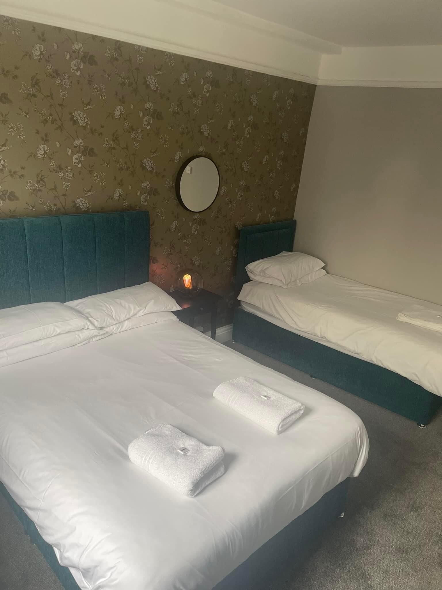 Une chambre d’hôtel avec deux lits, chacun agrémenté de draps et de serviettes blancs, un miroir rond au mur et un papier peint à motifs floraux.