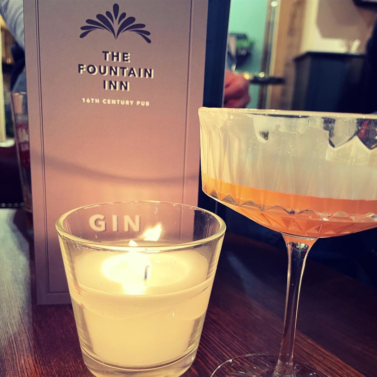 Ett ljus med etiketten ”GIN” och ett glas gin placerade framför en skylt med texten ”The Fountain Inn, en pub från 1500-talet”.