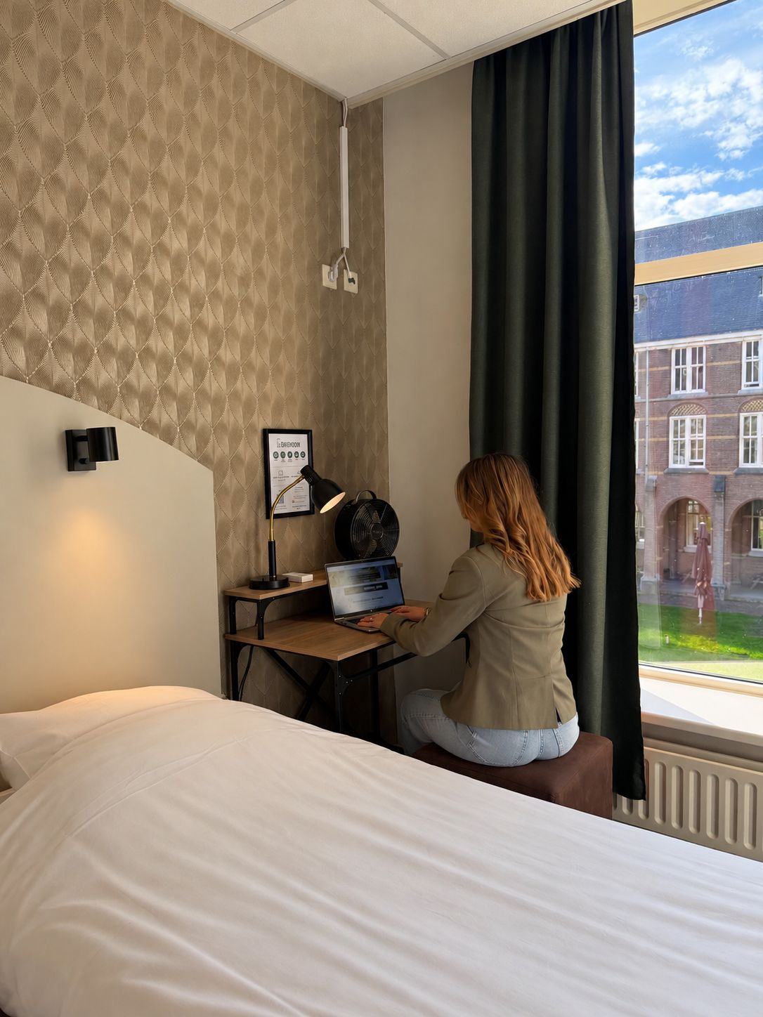 Une chambre d’hôtel avec un lit, un bureau avec un ordinateur portable, un ventilateur et une fenêtre donnant sur des immeubles.