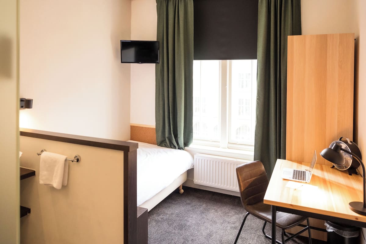 Une chambre d’hôtel avec un lit, un bureau avec un ordinateur portable, une chaise, un radiateur, un téléviseur fixé au mur et une fenêtre avec des rideaux.