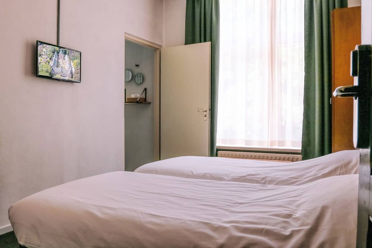Une chambre d’hôtel avec deux lits, un téléviseur fixé au mur, une porte donnant sur une salle de bain et une fenêtre avec des rideaux.