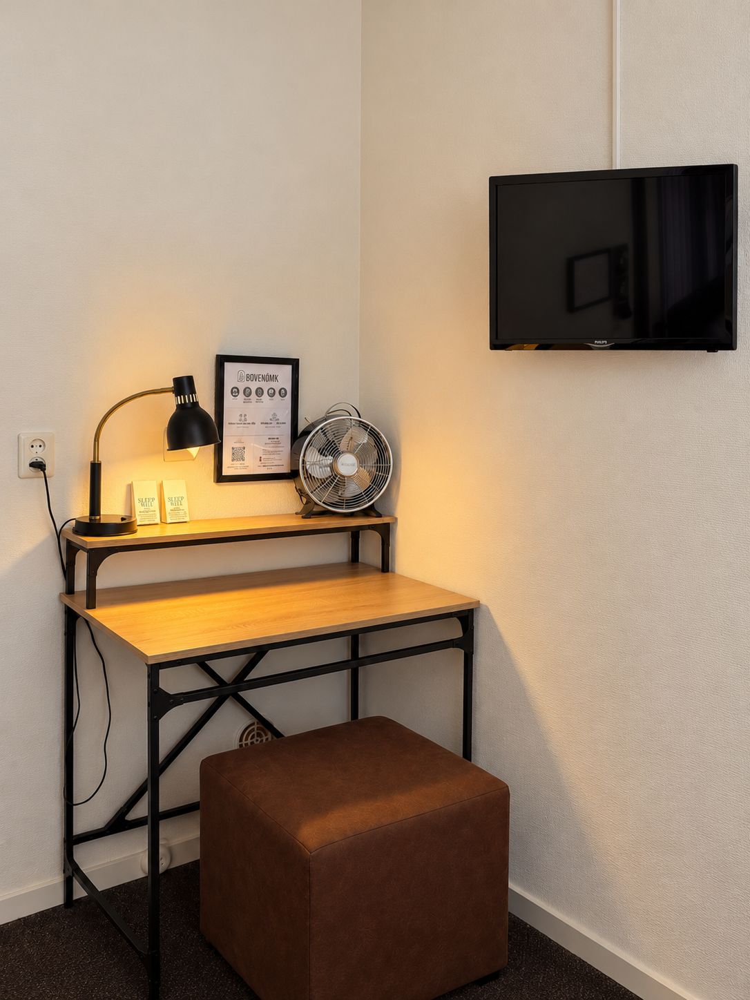 Um espaço de trabalho com uma escrivaninha, uma luminária, um ventilador, uma televisão e um banquinho marrom.