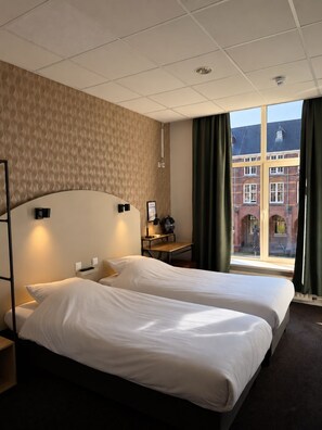 A hotel room with two beds, a desk, a chair, a window with curtains, and a view of buildings outside. -  Business & Eventlocation Bovendonk (Hoeven)