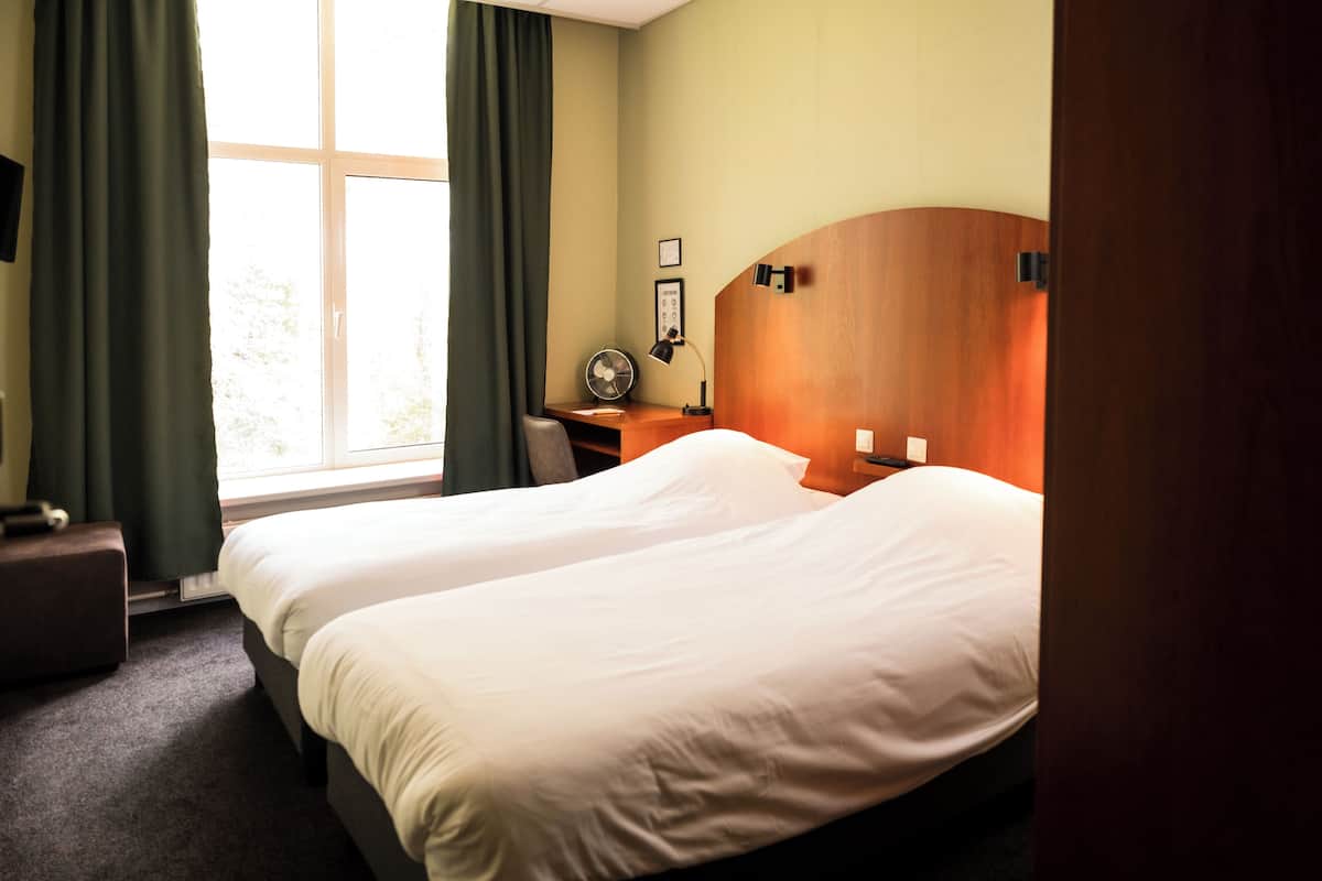 Une chambre d’hôtel avec deux lits, un bureau avec une lampe, un ventilateur et une fenêtre avec des rideaux.