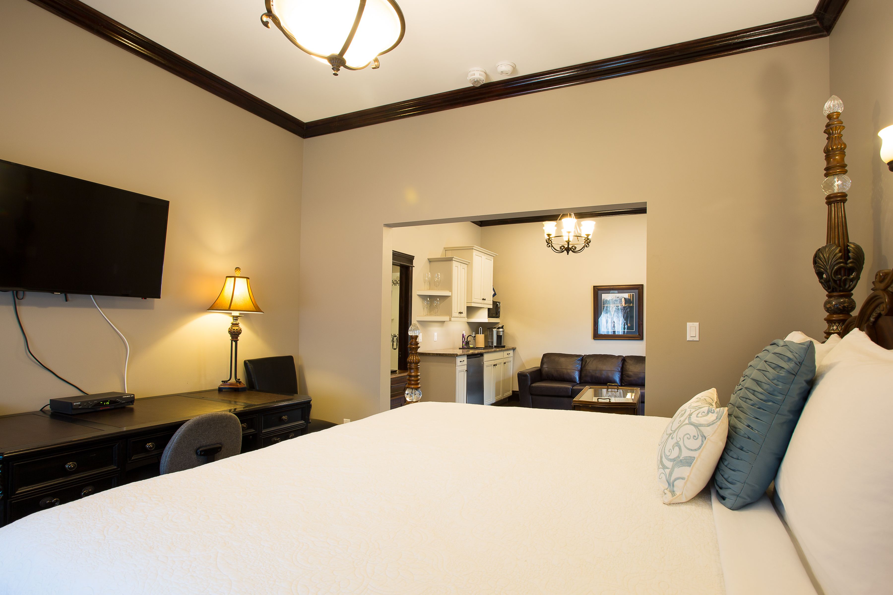 A hotel room with a large bed, a desk with a lamp, a television, and a kitchenette in the background.