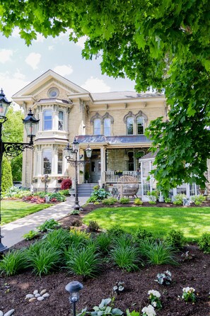 A large, historic building with a prominent bay window, a covered porch, and a well-maintained garden with various plants and a pathway leading to the entrance.