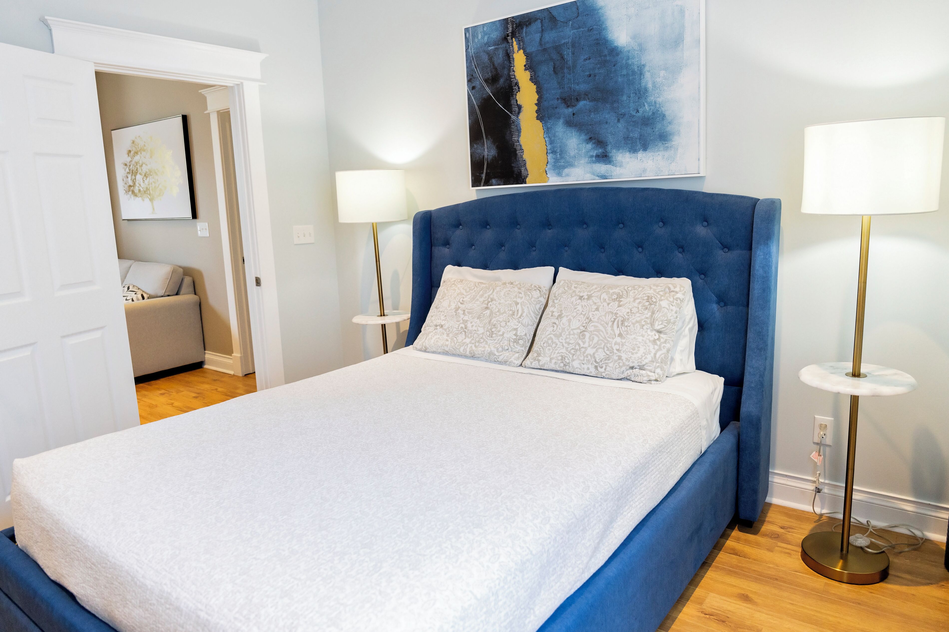 Een slaapkamer met een groot bed, twee bedlampen, een blauw hoofdeinde en een schilderij aan de muur.