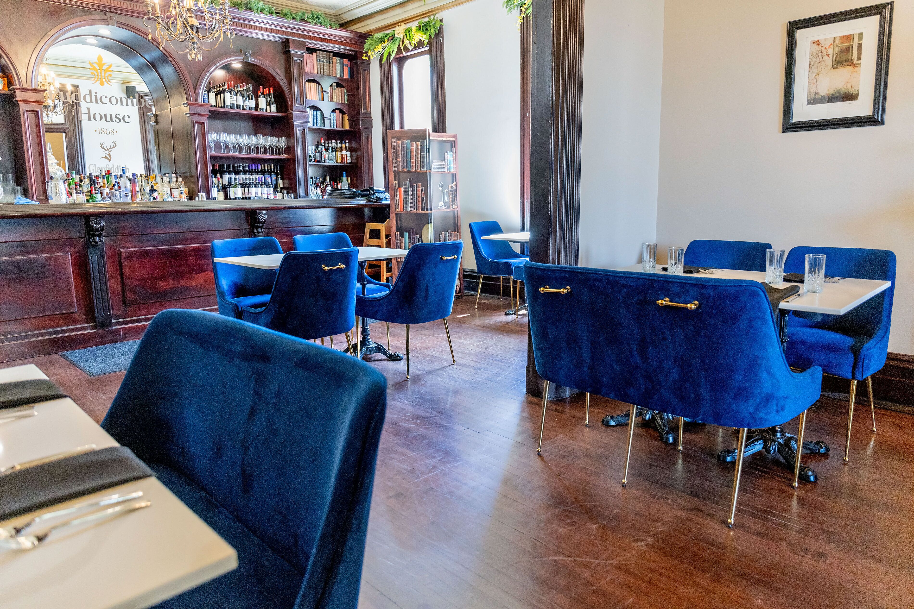 Un restaurante con sillas de terciopelo azul, una zona de bar con estantería, y un cartel que dice "Ridicom House 1868".