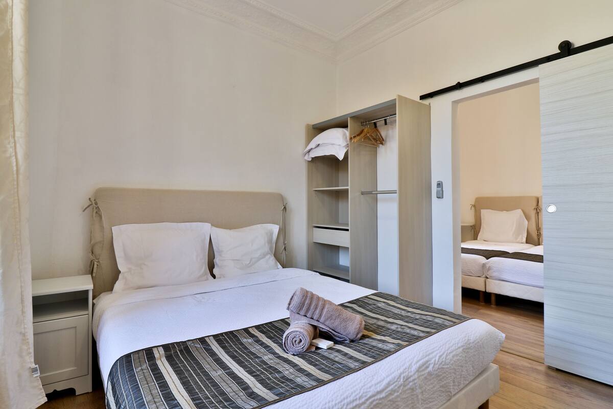 A neatly made bed with a striped bedspread, a bedside table, and an open wardrobe with hanging clothes.