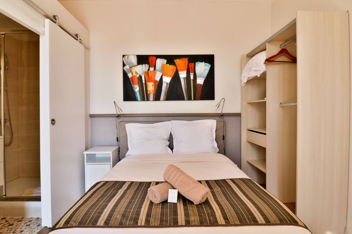 A hotel room with a bed, a painting of paintbrushes, a towel, and a walk-in closet.