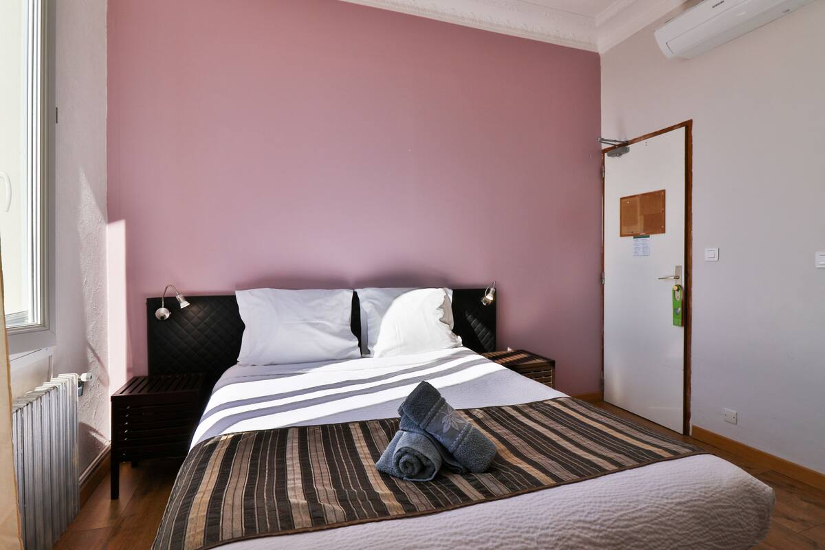 A bedroom with a bed, a nightstand, a radiator, and a door with a bulletin board.