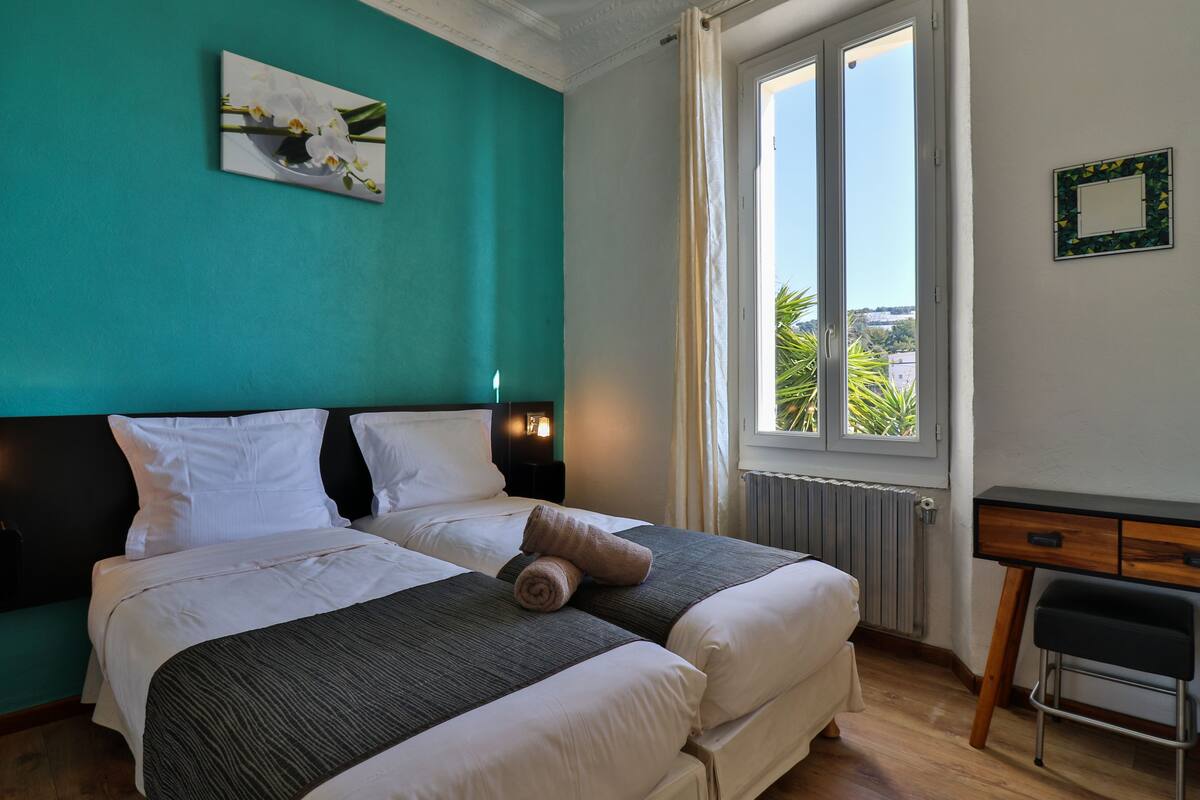 A double bed with white linens and a dark grey blanket, a small desk with a chair, a window with a view of greenery, and a framed picture on the wall.