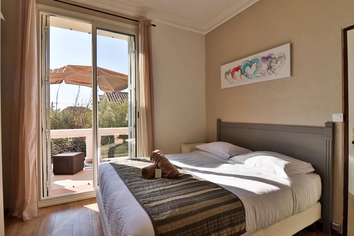 A bedroom with a bed, a window with curtains, a balcony with an umbrella, and a painting on the wall.