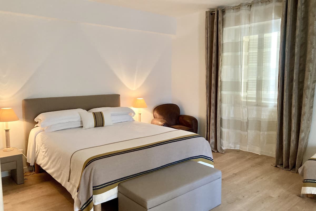Una cama bien tendida con cabecera, una mesita de noche con lámpara, una silla, una ventana con cortinas y piso de madera.