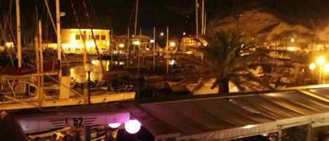 A marina at night with boats docked, a covered area with people, and buildings in the background.