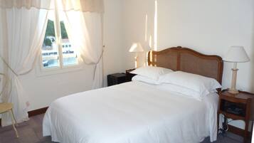 A neatly made bed in a hotel room with a wooden headboard, a nightstand with a lamp, and a chair by the window.
