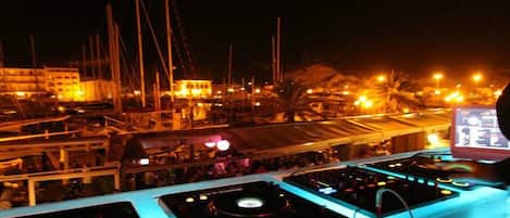 A DJ setup with multiple turntables and mixers, overlooking a marina with boats and a building in the background.