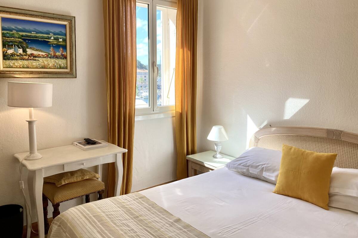 Habitación de hotel con cama, escritorio con lámpara, mesita de noche con lámpara, un cuadro en la pared y una ventana con cortinas.