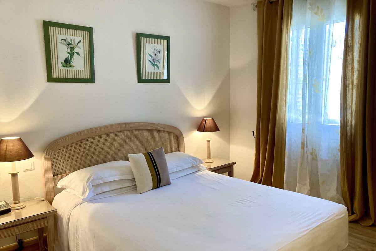 Una cama bien hecha con ropa de cama blanca, dos cuadros florales enmarcados en la pared, una lámpara de noche y una ventana con cortinas.
