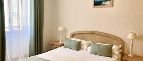 A neatly arranged hotel room with a large bed, two bedside tables with lamps, a painting on the wall, and a view of the sea through the window.