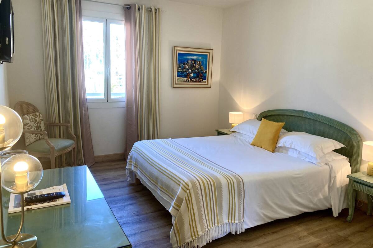 Una habitación de hotel con cama, una silla, una mesa con lámpara, un cuadro en la pared y una ventana con cortinas.