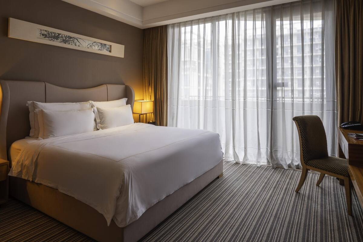 A hotel room with a large bed, a desk with a chair, a lamp, and a window with sheer curtains.